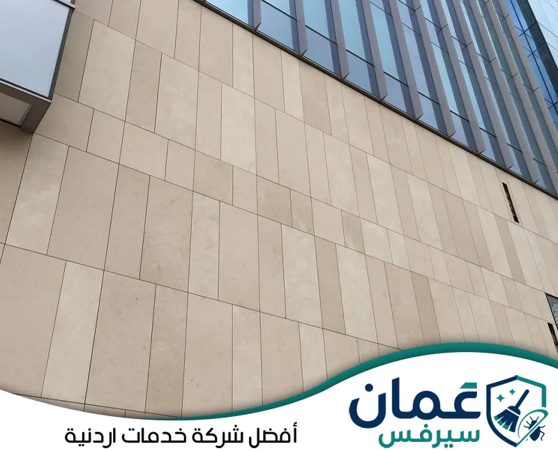 احجز تنظيف واجهات الكلادينج في الأردن مع عمان سيرفس على 0798779667. عامل يستخدم معدات الحبال والضغط المائي لتنظيف واجهة كلادينج مرتفعة وإزالة الأتربة والبقع من الألواح الخارجية.