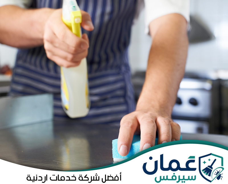 احجز تنظيف المطبخ التجاري مع عمان سيرفس واتصل على 0798779667. عامل ينظف سطحًا معدنيًا داخل مطبخ تجاري باستخدام منظف وقطعة إسفنج لإزالة الأوساخ.