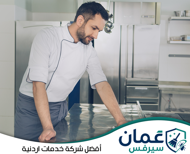 تواصل مع شركة تنظيف المطاعم في الأردن على 0798779667. طاهٍ داخل مطبخ تجاري يقف أمام سطح تحضير معدني نظيف مع معدات وثلاجات في الخلفية.
