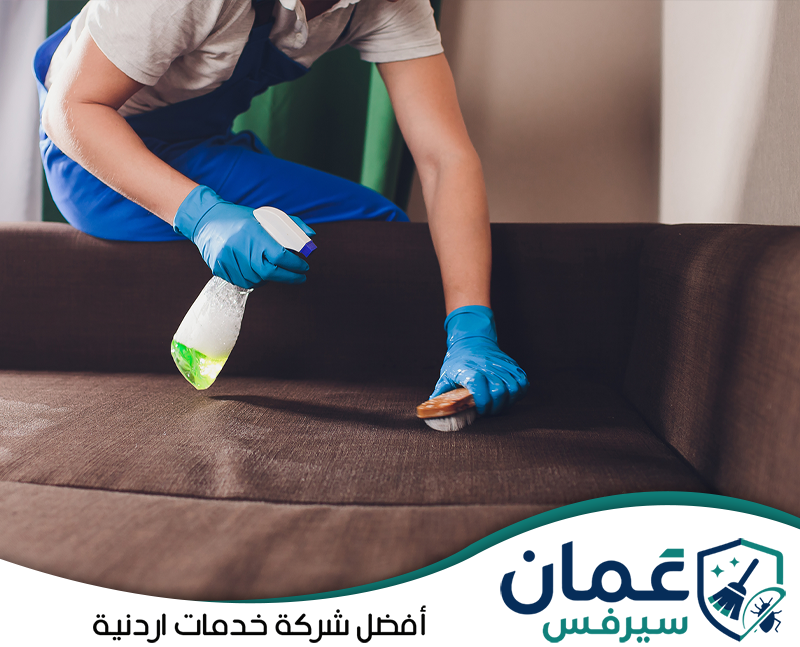 تواصل مع عمان سيرفس لتنظيف بقع الكنب على 0798779667. عامل يعالج سطح كنب بني باستخدام محلول تنظيف وفرشاة يدوية للوصول إلى البقع والأوساخ.