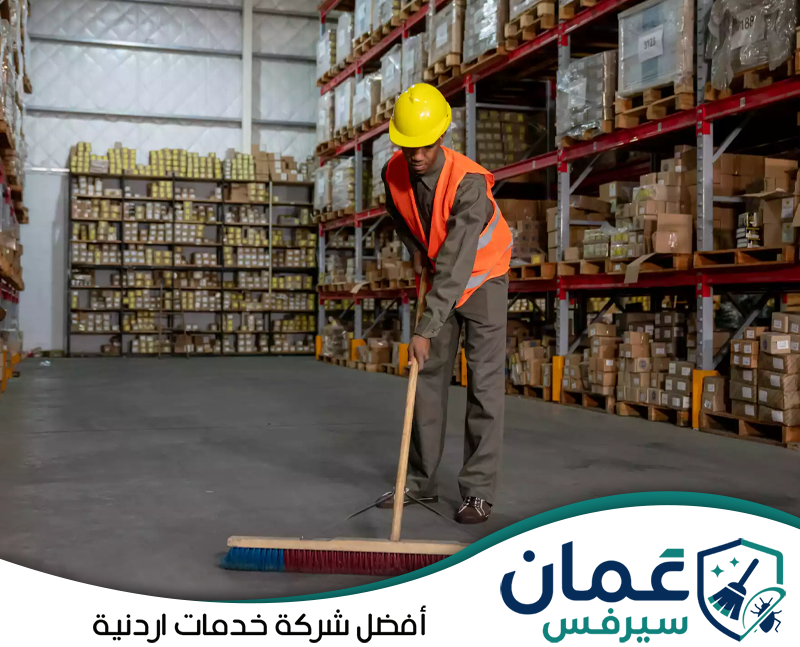 اتصل على 0798779667 لطلب خدمة تنظيف مستودعك. عامل يرتدي معدات السلامة أثناء كنس وتنظيف أرضية مستودع واسع بين الأرفف والبضائع المخزنة.